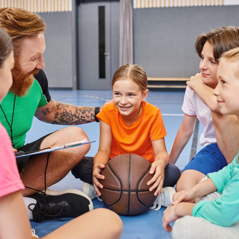 A man teacher with a diverse group of kids sitting around a basketball, engaging in a lively lesson in a bright classroom setting. A man teacher with a diverse group of kids sitting around a basketball, engaging in a lively lesson in a bright classroom setting.